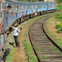 Un tren azul de Sri Lanka avanza por las vías, con pasajeros asomados a las ventanas y otros viajando en los estribos.