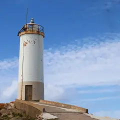 Faro blanco sobre rocas, parte de la ruta de los faros Galicia en coche. Cielo azul con nubes.