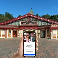 Mano sujetando un teléfono que muestra la opción "Cola virtual" para conocer a Mickey Mouse en Disneyland Paris, frente al edificio "Meet Mickey Mouse".