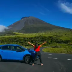 Mujer con los brazos abiertos junto a un coche azul, con el imponente volcán de la isla del Pico de fondo.