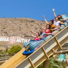 Grupo de amigos ríe y grita en una atracción del parque temático Terra Mítica, con el logo del parque al fondo.