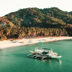 Barco blanco en aguas turquesas, playa de arena blanca con palmeras y colinas verdes. Un destino soñado para tu viaje a Filipinas, ¡el precio vale la pena!