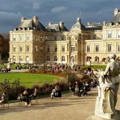Gente paseando y relajándose en los jardines de Luxemburgo, con el palacio de fondo y una estatua en primer plano.