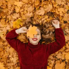 Mujer sonríe acostada sobre hojas secas, cubriendo un ojo con una hoja de arce. ¡Perfectas vacaciones de otoño!