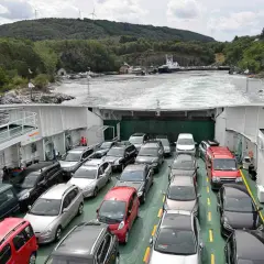 Coches en ferry cruzando un fiordo en Noruega. Paisaje verde y montañoso con molinos de viento.