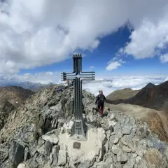 Cima de montaña con cruz metálica. Una persona camina por la ruta fácil, rodeada de picos y nubes.