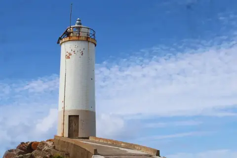 Faro blanco sobre rocas, parte de la ruta de los faros Galicia en coche. Cielo azul con nubes.