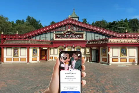 Mano sujetando un teléfono que muestra la opción "Cola virtual" para conocer a Mickey Mouse en Disneyland Paris, frente al edificio "Meet Mickey Mouse".