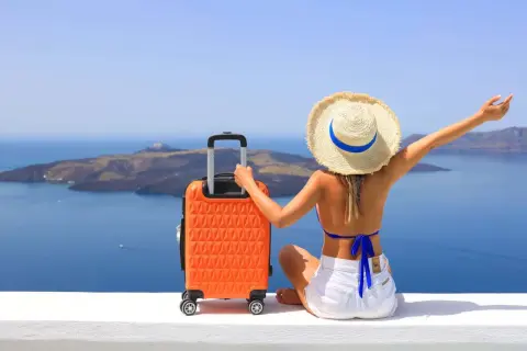 Mujer con sombrero y maleta naranja, lista para decidir dónde viajar en agosto, con vistas al mar y a una isla.