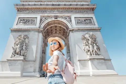 Una mujer con sombrero de paja y mochila rosa extiende la mano frente al Arco del Triunfo, un ícono de las cosas típicas de Francia.