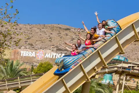 Grupo de amigos ríe y grita en una atracción del parque temático Terra Mítica, con el logo del parque al fondo.