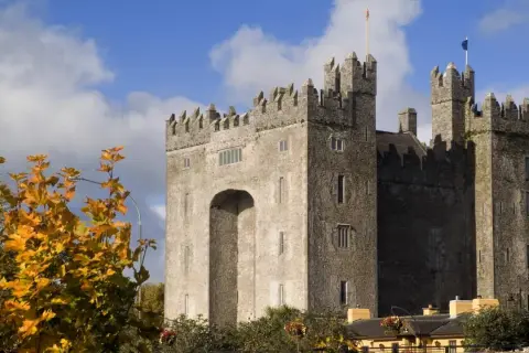 El imponente castillo de Bunratty se alza bajo un cielo azul con nubes, rodeado de follaje otoñal.