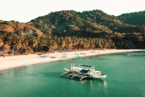 Barco blanco en aguas turquesas, playa de arena blanca con palmeras y colinas verdes. Un destino soñado para tu viaje a Filipinas, ¡el precio vale la pena!