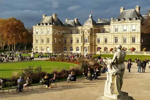 Gente paseando y relajándose en los jardines de Luxemburgo, con el palacio de fondo y una estatua en primer plano.
