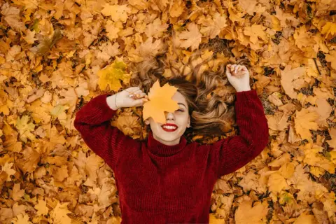 Mujer sonríe acostada sobre hojas secas, cubriendo un ojo con una hoja de arce. ¡Perfectas vacaciones de otoño!