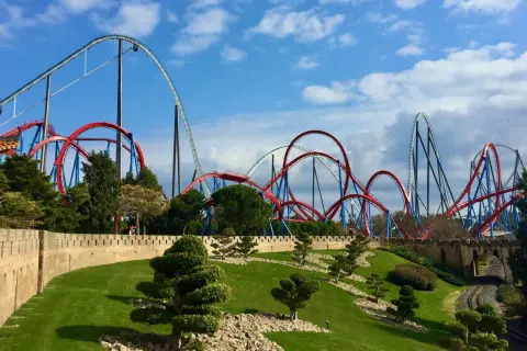 Montañas rusas rojas y azules se elevan sobre un paisaje verde y una muralla de piedra. ¡Qué ganas de ir a PortAventura!