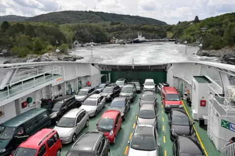 Coches en ferry cruzando un fiordo en Noruega. Paisaje verde y montañoso con molinos de viento.