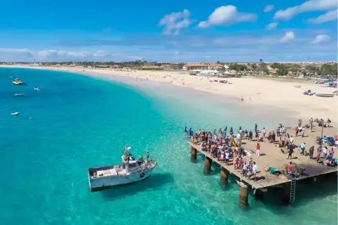 Turistas en un muelle de madera en Cabo Verde, con aguas turquesas y una playa de arena. El turismo en Cabo Verde puede ser peligroso si no se toman precauciones.