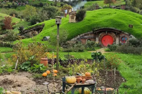 Un jardín exuberante en Hobbiton, Nueva Zelanda, con calabazas y flores, y una casa hobbit con puerta roja.