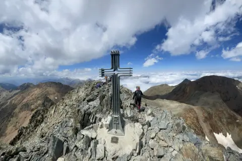 Cima de montaña con cruz metálica. Una persona camina por la ruta fácil, rodeada de picos y nubes.