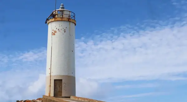 Faro blanco sobre rocas, parte de la ruta de los faros Galicia en coche. Cielo azul con nubes.