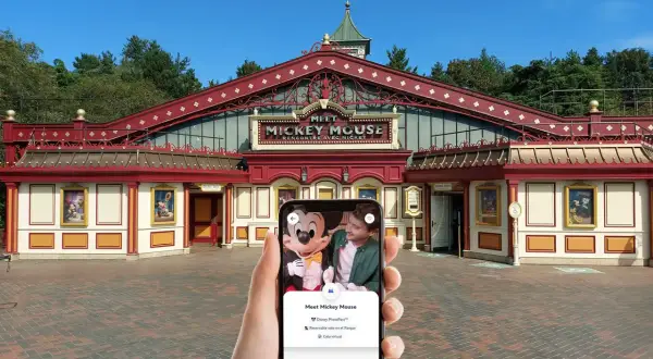 Mano sujetando un teléfono que muestra la opción "Cola virtual" para conocer a Mickey Mouse en Disneyland Paris, frente al edificio "Meet Mickey Mouse".