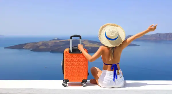 Mujer con sombrero y maleta naranja, lista para decidir dónde viajar en agosto, con vistas al mar y a una isla.