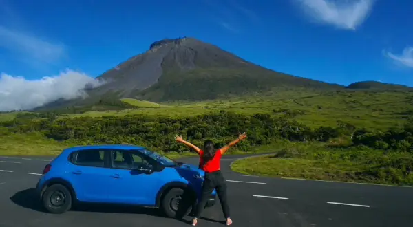 Mujer con los brazos abiertos junto a un coche azul, con el imponente volcán de la isla del Pico de fondo.