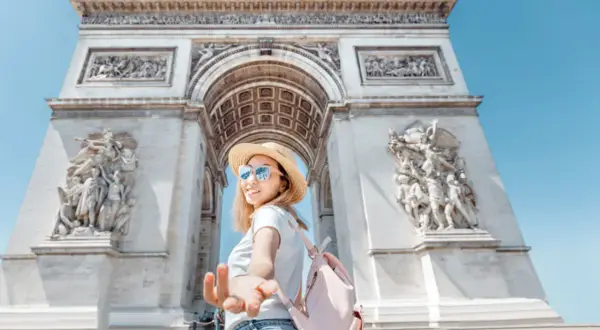 Una mujer con sombrero de paja y mochila rosa extiende la mano frente al Arco del Triunfo, un ícono de las cosas típicas de Francia.