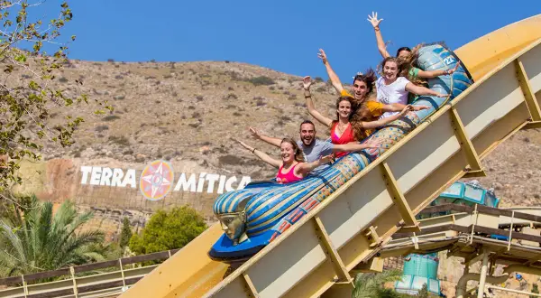Grupo de amigos ríe y grita en una atracción del parque temático Terra Mítica, con el logo del parque al fondo.