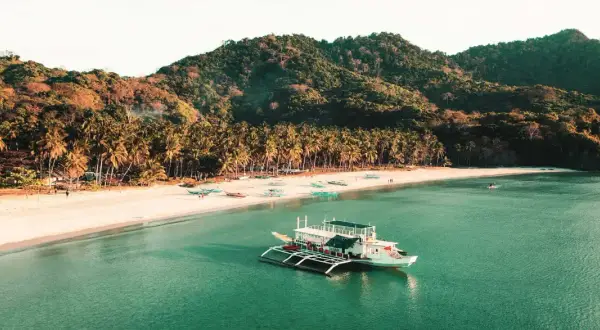 Barco blanco en aguas turquesas, playa de arena blanca con palmeras y colinas verdes. Un destino soñado para tu viaje a Filipinas, ¡el precio vale la pena!