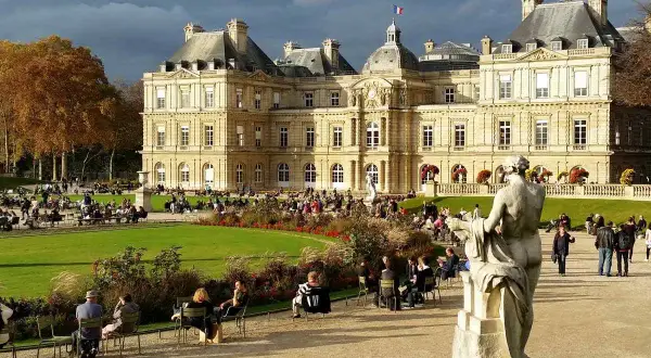 Gente paseando y relajándose en los jardines de Luxemburgo, con el palacio de fondo y una estatua en primer plano.