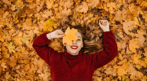 Mujer sonríe acostada sobre hojas secas, cubriendo un ojo con una hoja de arce. ¡Perfectas vacaciones de otoño!