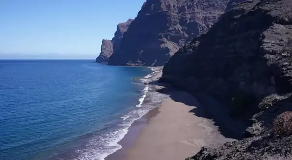 Costa salvaje con acantilados oscuros y una playa de arena dorada bañada por el mar azul. Un rincón secreto entre las mejores playas de Gran Canaria.