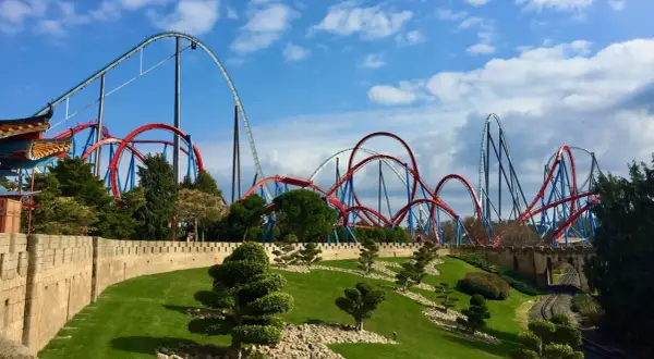Montañas rusas rojas y azules se elevan sobre un paisaje verde y una muralla de piedra. ¡Qué ganas de ir a PortAventura!