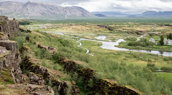 Paisaje de Thingvellir National Park con un río serpenteante, montañas al fondo y una grieta rocosa en primer plano.