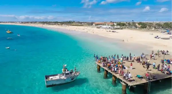 Turistas en un muelle de madera en Cabo Verde, con aguas turquesas y una playa de arena. El turismo en Cabo Verde puede ser peligroso si no se toman precauciones.