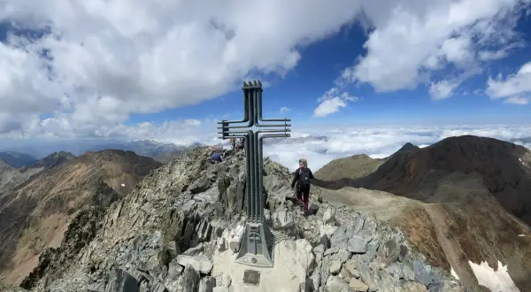 Cima de montaña con cruz metálica. Una persona camina por la ruta fácil, rodeada de picos y nubes.