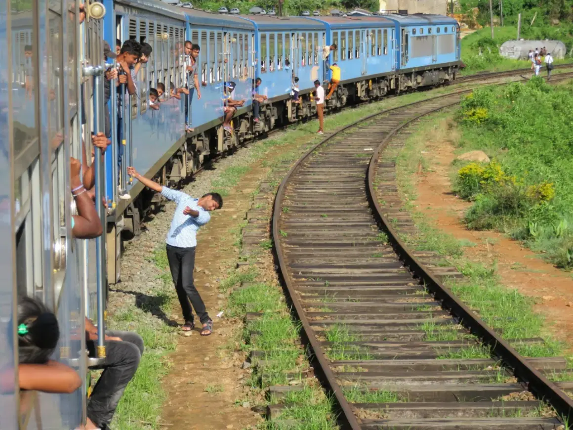 Un tren azul de Sri Lanka avanza por las vías, con pasajeros asomados a las ventanas y otros viajando en los estribos.