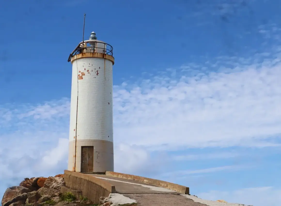 Faro blanco sobre rocas, parte de la ruta de los faros Galicia en coche. Cielo azul con nubes.