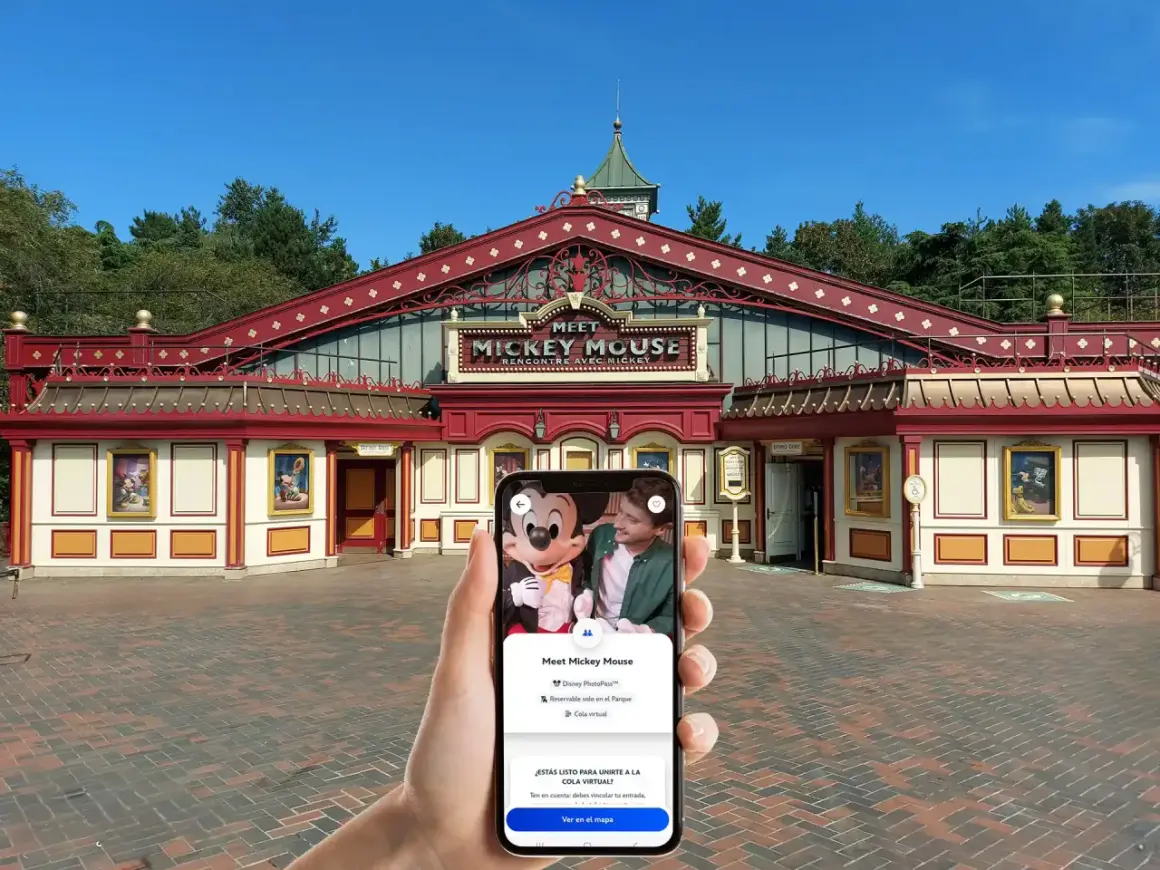 Mano sujetando un teléfono que muestra la opción "Cola virtual" para conocer a Mickey Mouse en Disneyland Paris, frente al edificio "Meet Mickey Mouse".