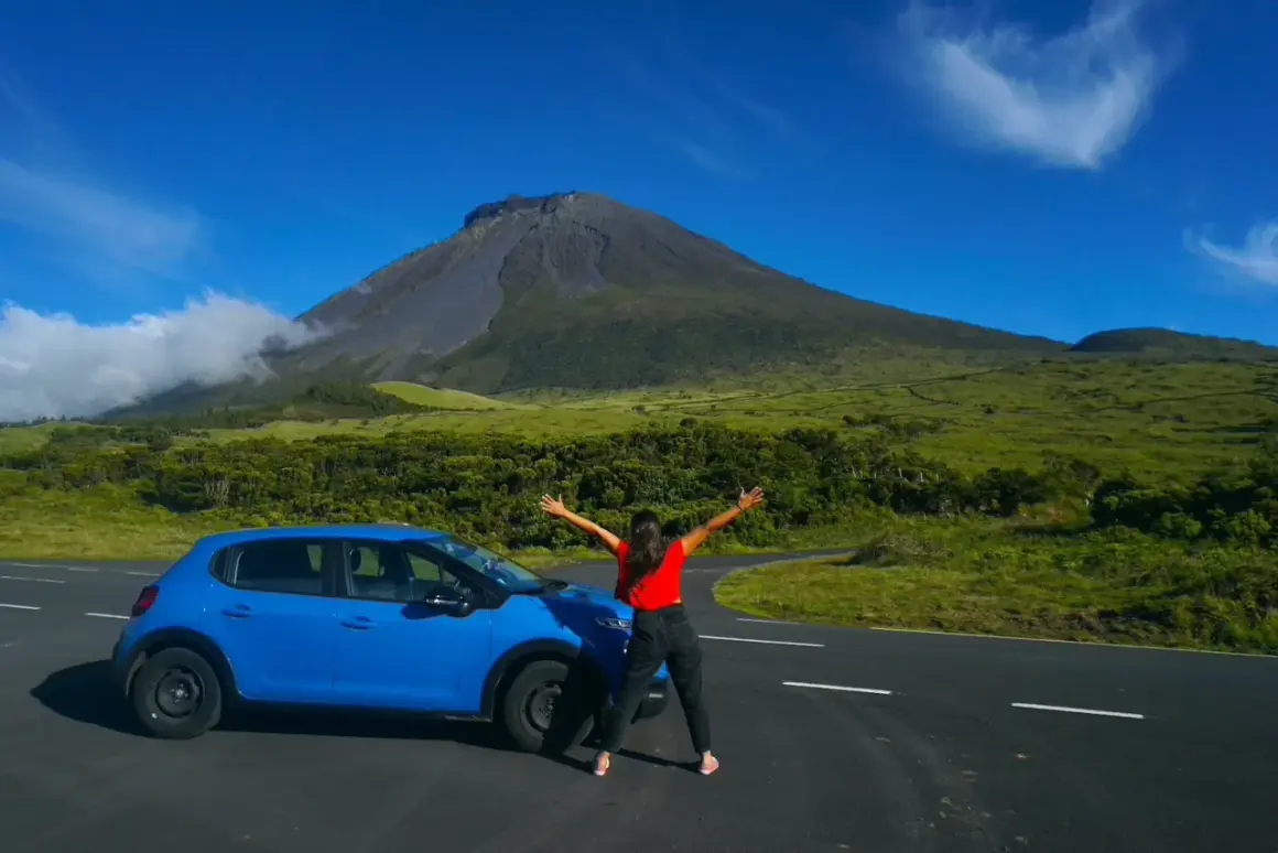 Mujer con los brazos abiertos junto a un coche azul, con el imponente volcán de la isla del Pico de fondo.