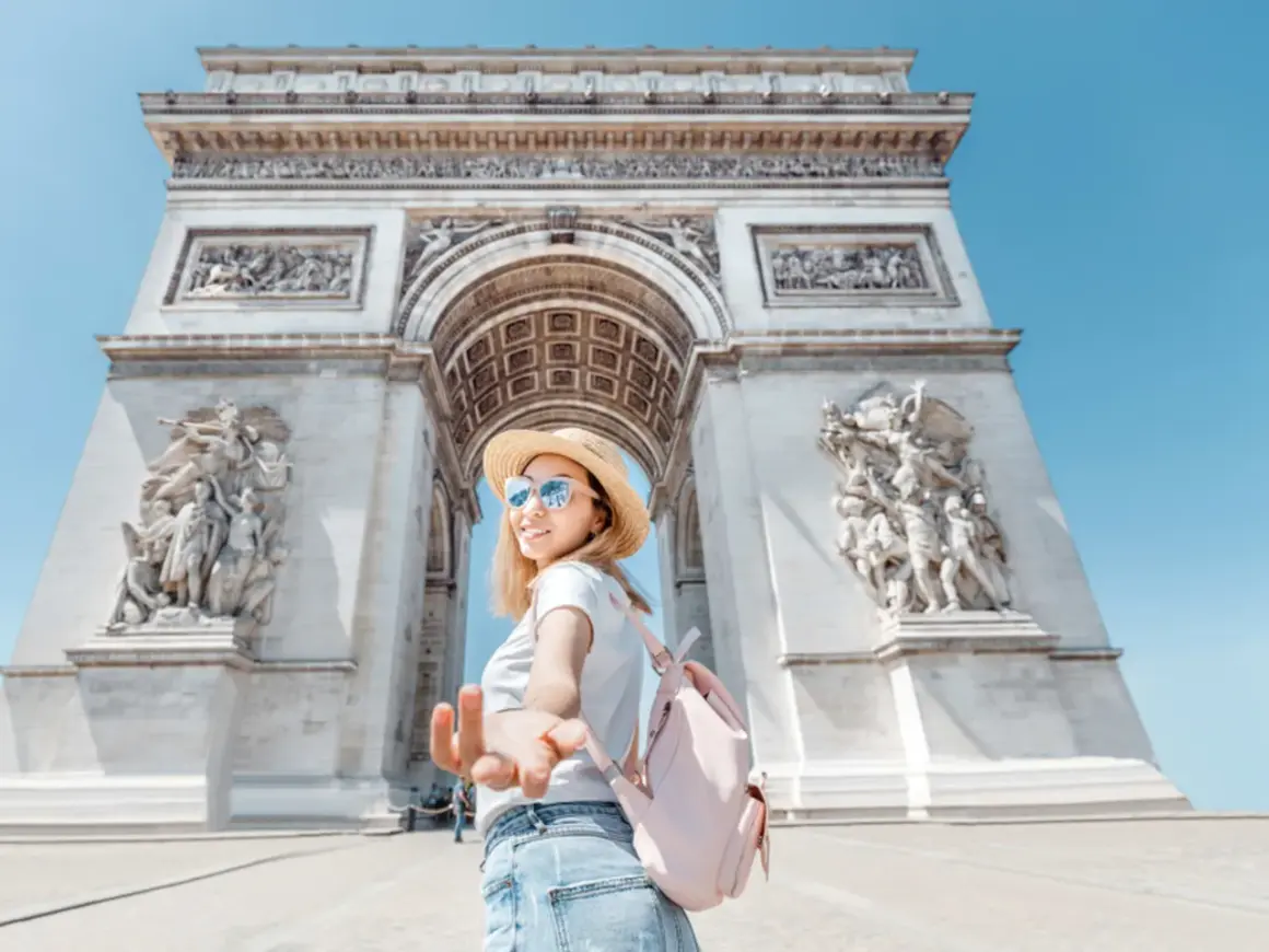 Una mujer con sombrero de paja y mochila rosa extiende la mano frente al Arco del Triunfo, un ícono de las cosas típicas de Francia.