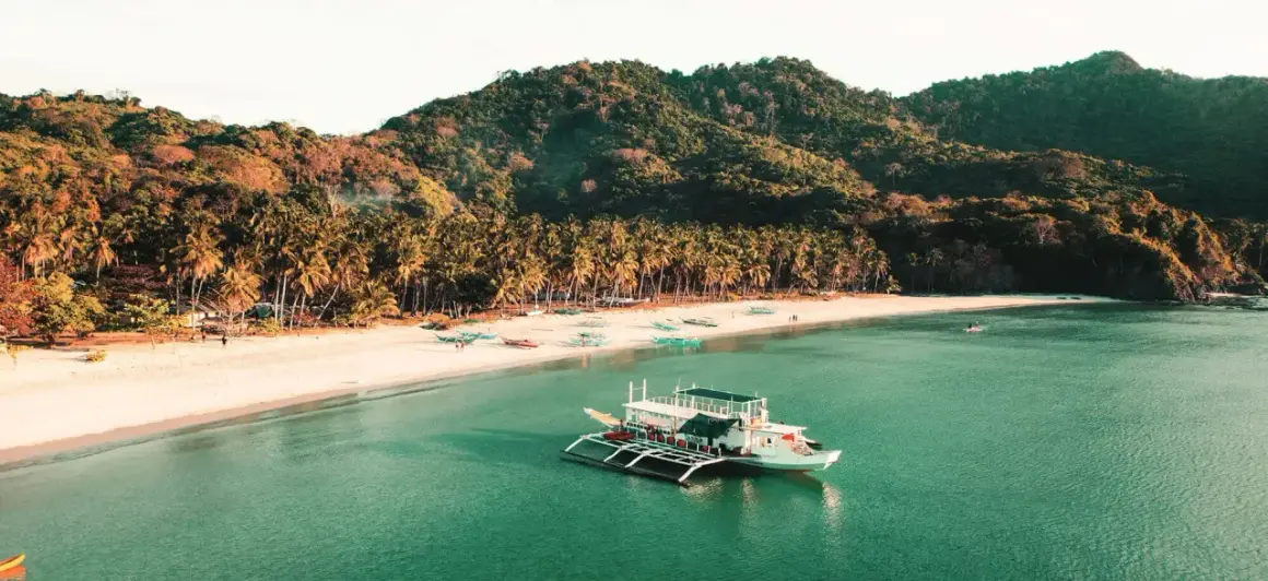 Barco blanco en aguas turquesas, playa de arena blanca con palmeras y colinas verdes. Un destino soñado para tu viaje a Filipinas, ¡el precio vale la pena!