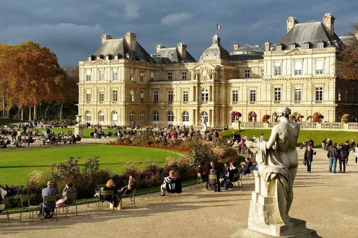 Gente paseando y relajándose en los jardines de Luxemburgo, con el palacio de fondo y una estatua en primer plano.