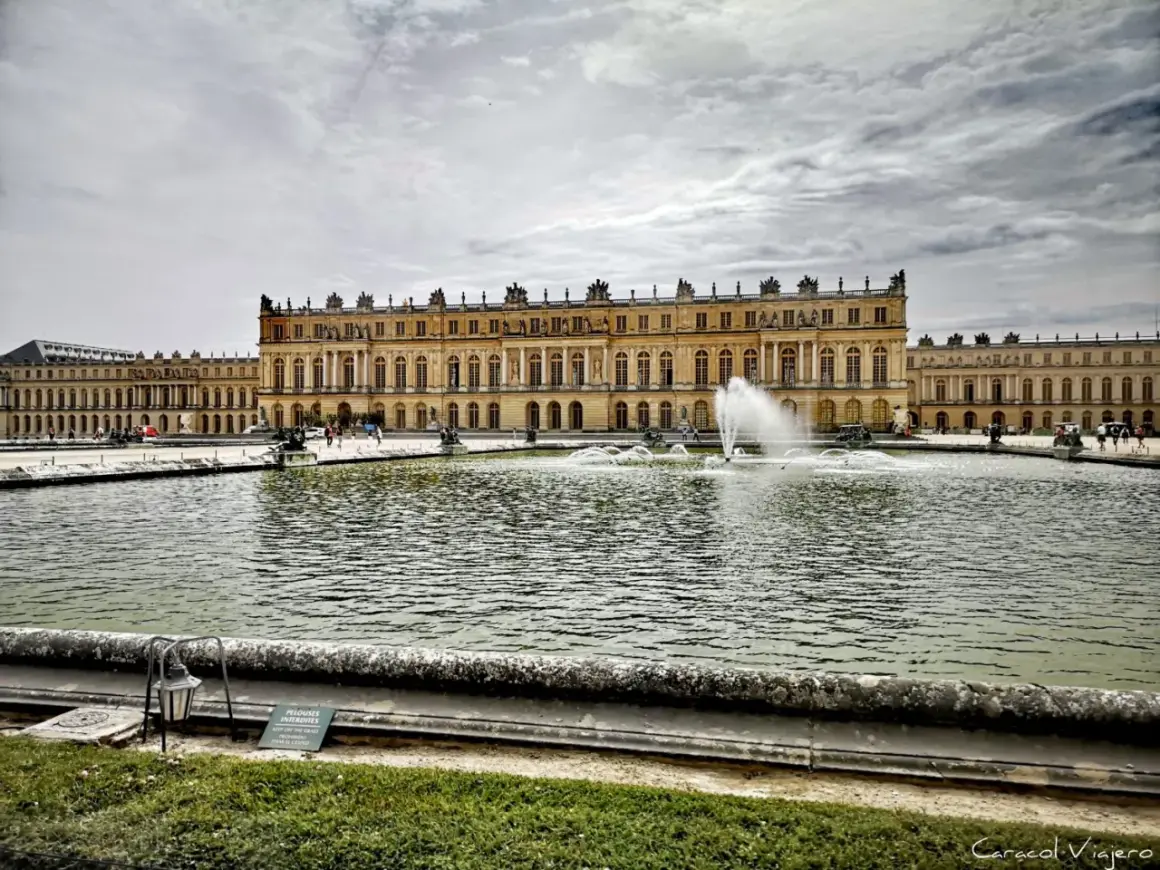 El **Palacio de Versalles** se alza majestuoso con sus fuentes danzantes y un estanque reflejando el cielo nublado.