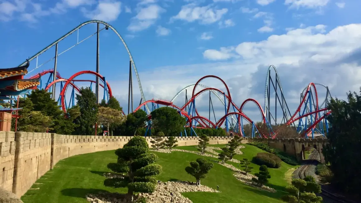 Montañas rusas rojas y azules se elevan sobre un paisaje verde y una muralla de piedra. ¡Qué ganas de ir a PortAventura!