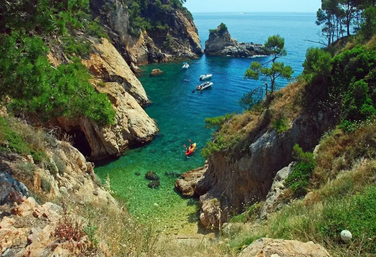 Un kayak rojo navega en aguas turquesas de una cala rocosa, ideal para saber donde viajar en junio.