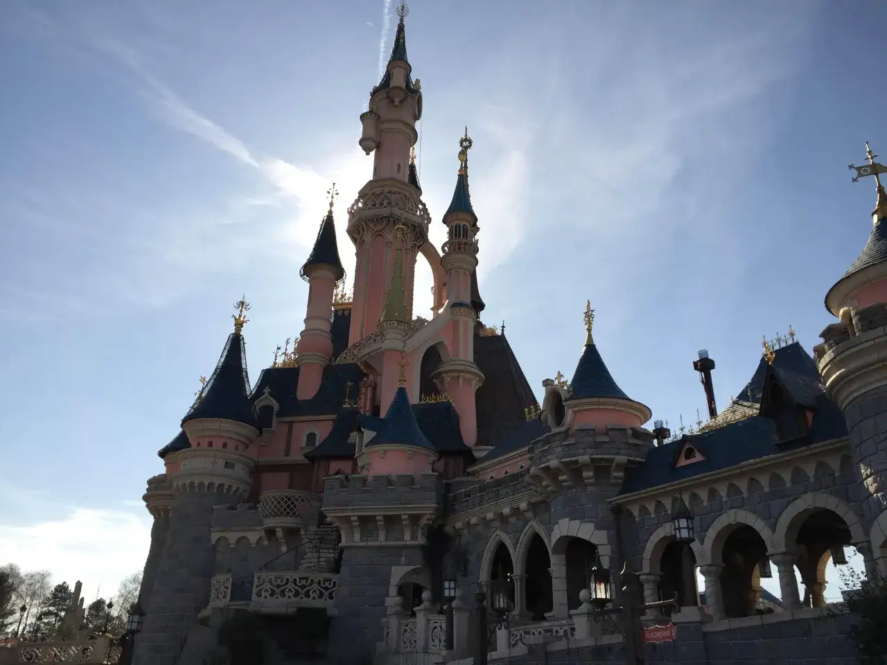 El castillo de la Bella Durmiente se alza majestuoso bajo un cielo azul con nubes tenues. Sus torres rosadas y tejados azules brillan, invitando a un cuento de hadas.