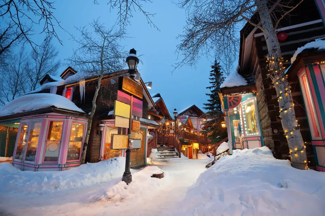 Encantador pueblo nevado, ideal para tus destinos para viajar en enero. Luces c&aacute;lidas iluminan las tiendas y la nieve cubre el camino.